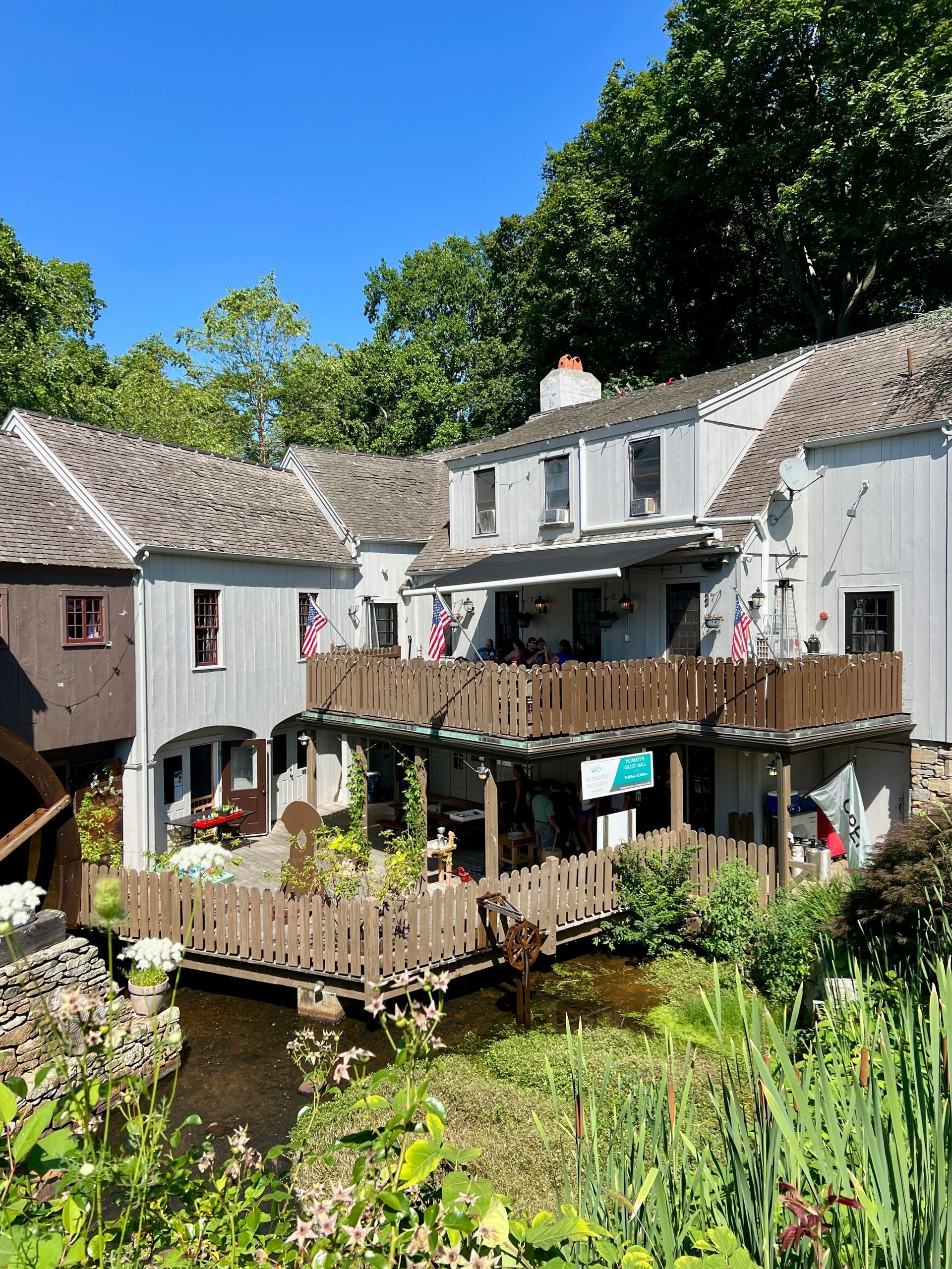 A white wooden building with a wooden deck overlooks a small stream, surrounded by lush green trees on a sunny day.