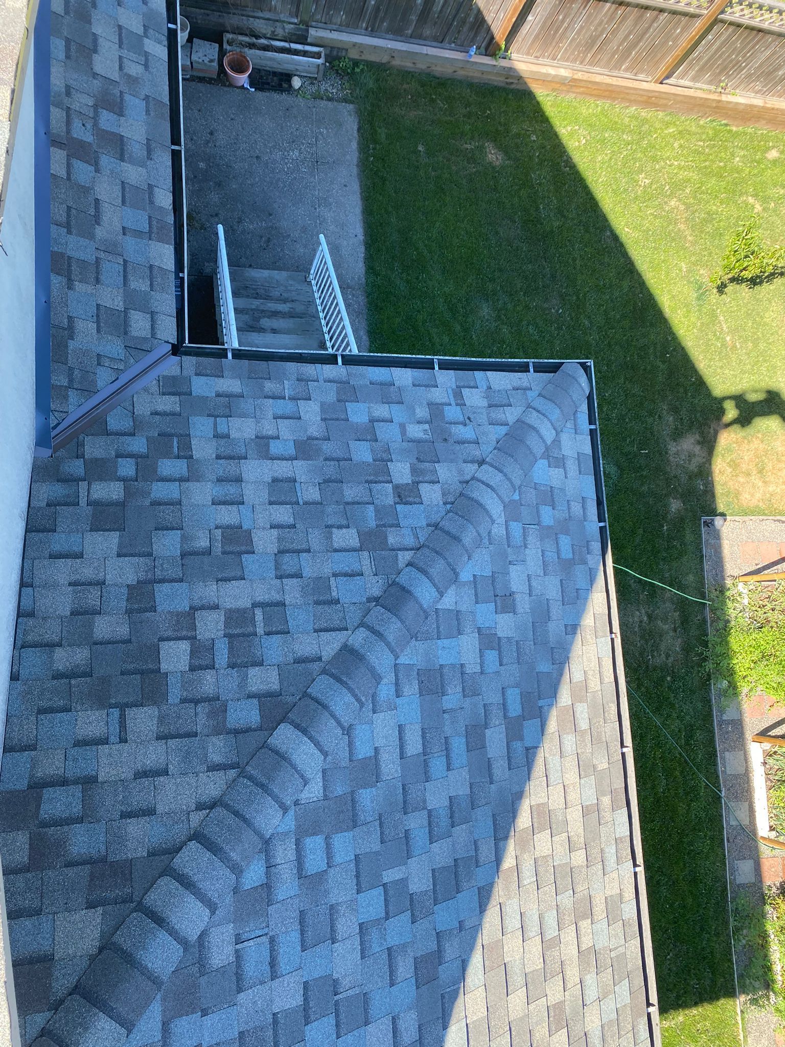 High-angle view of a grey shingled roof meeting at a corner, overlooking a green backyard and patio area.