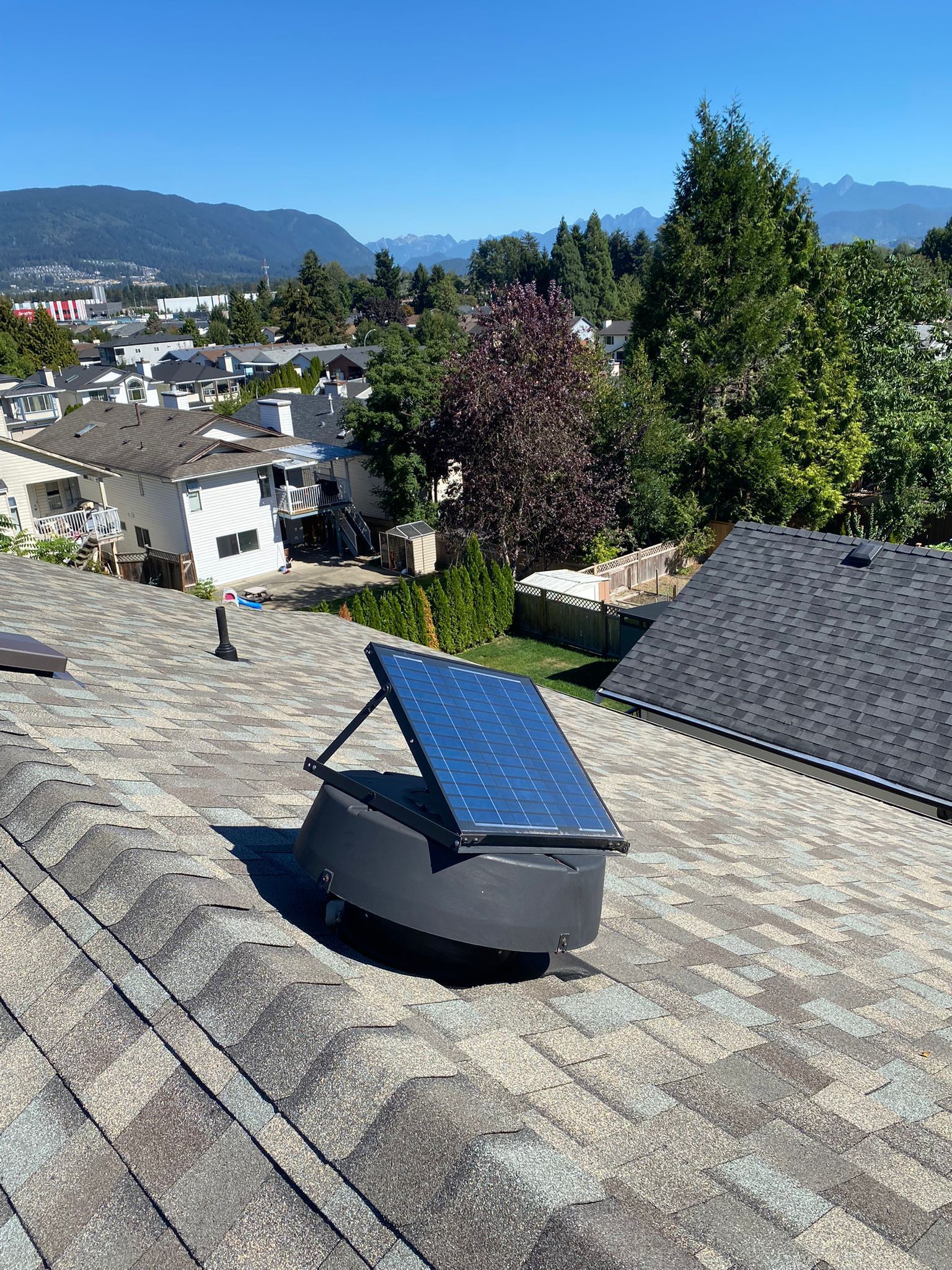 A solar-powered roof attic vent installed on a shingled residential roof against a background of trees and mountains.