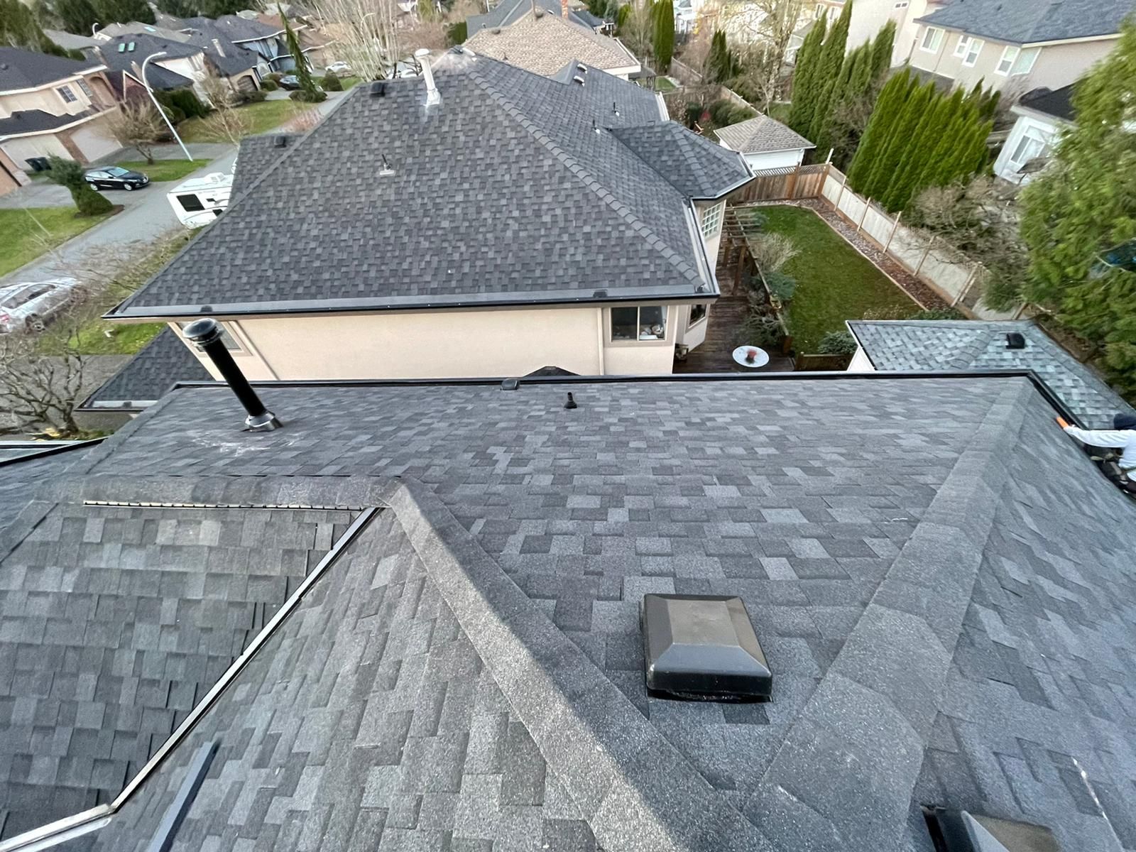 An elevated view of grey shingled roofs on a suburban house, featuring a central vent and nearby residential homes.