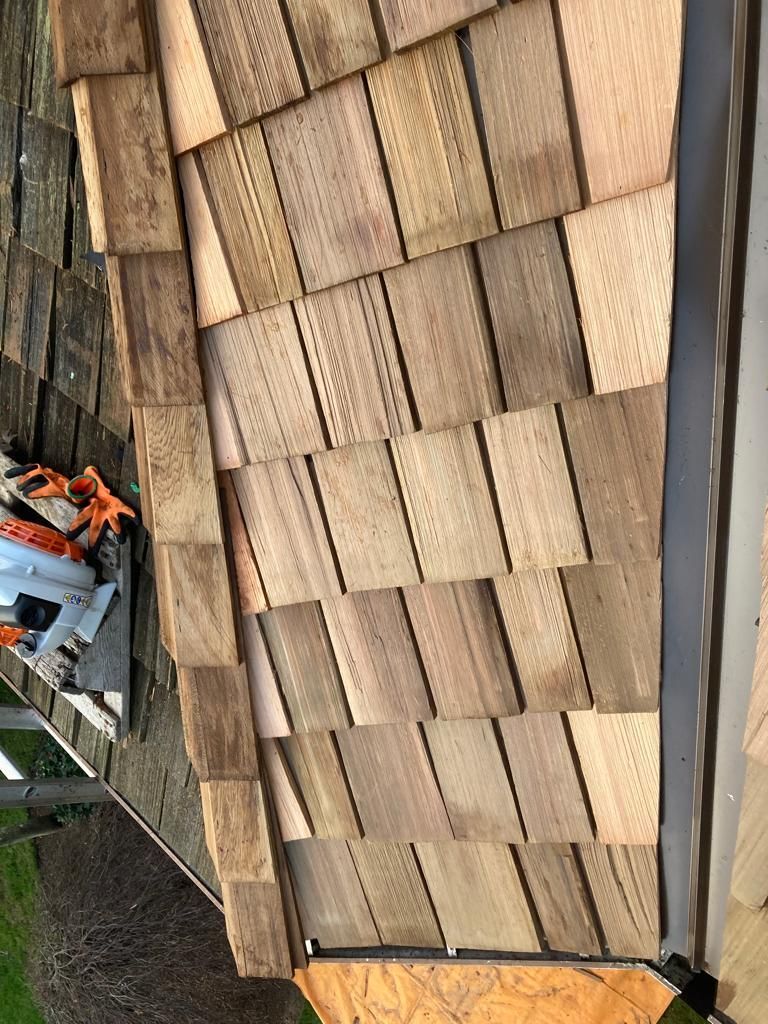 A close-up of a residential roof under construction featuring new, light brown wood shakes installed along the edge.