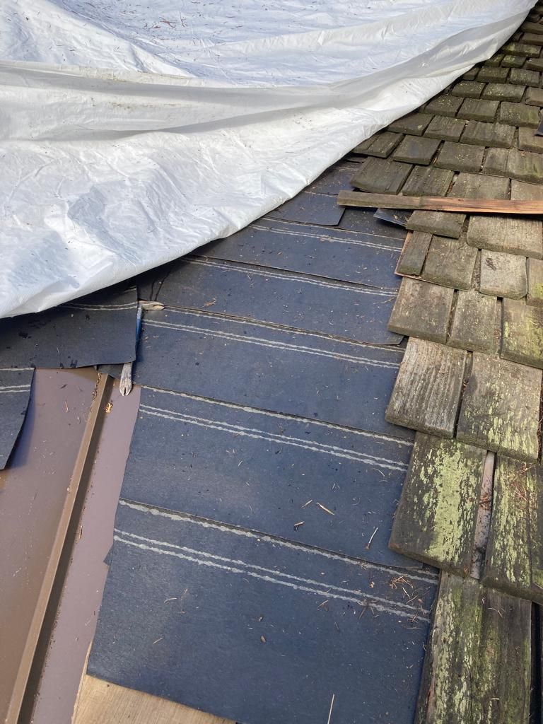 A white tarp covering a section of a roof where shingles have been removed, exposing the dark underlayment and flashing.