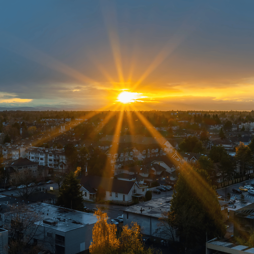 A radiant sun setting over a residential neighborhood, casting long, golden sunburst rays across the rooftops.
