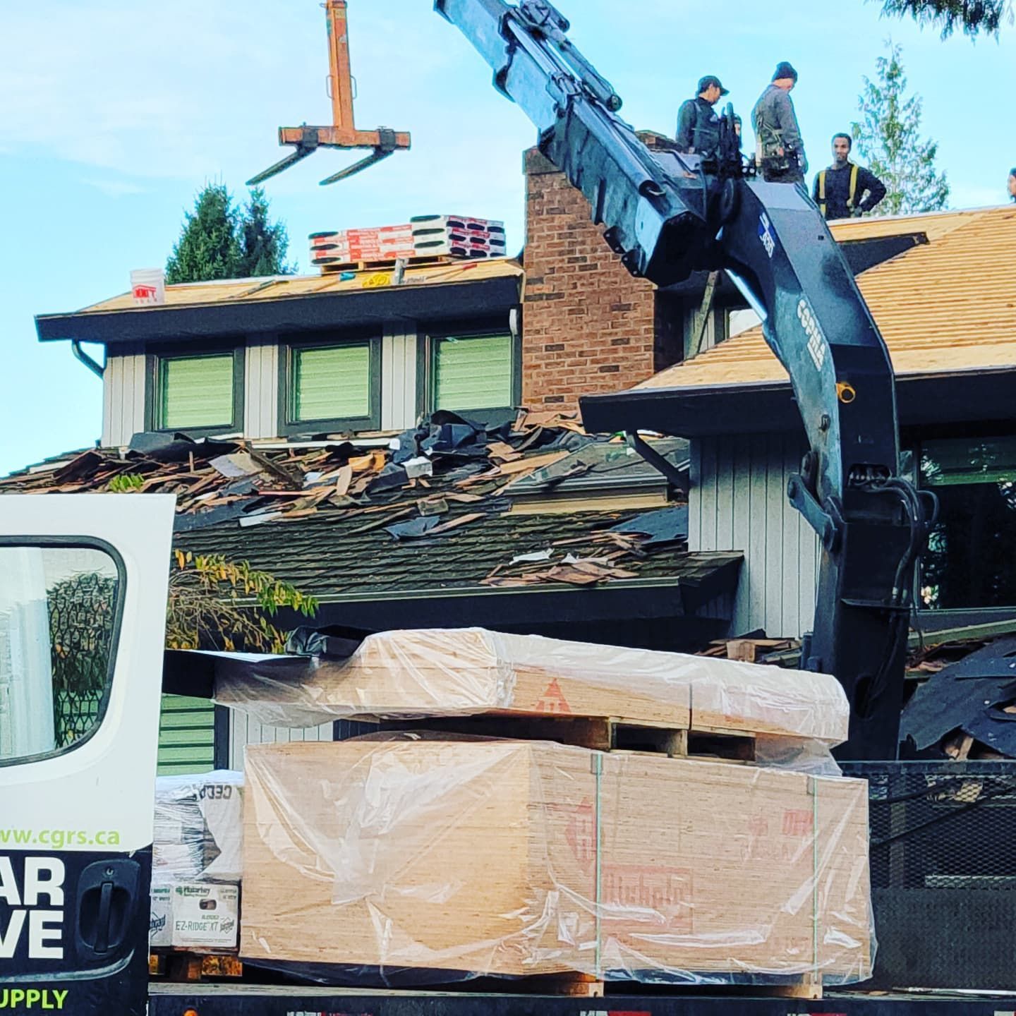 A crane lifts roofing supplies to workers on a residential house undergoing a roof replacement.