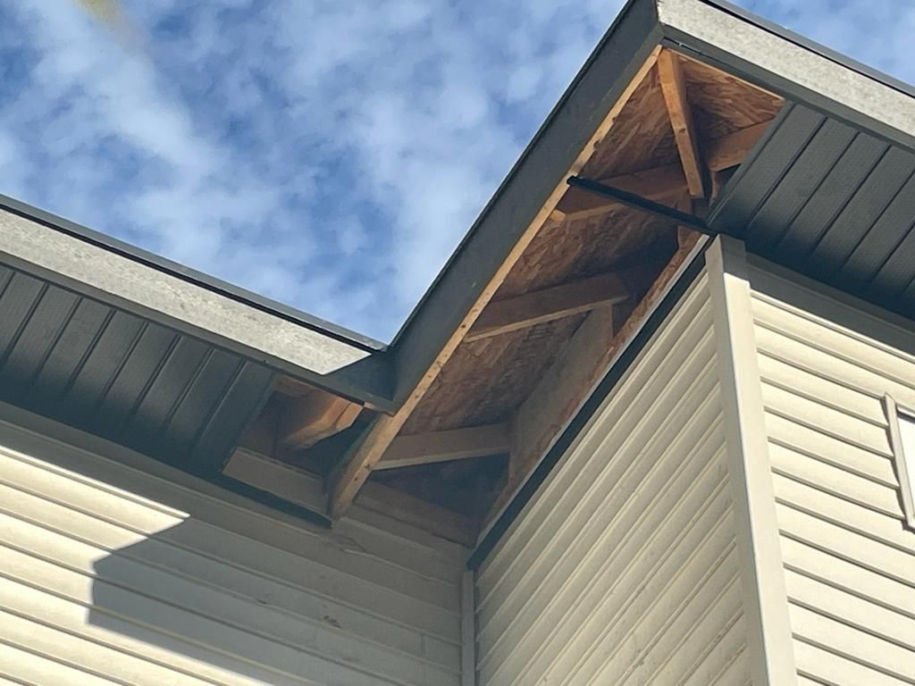 House roof corner with cream siding, dark trim, and exposed wood rafters against a blue sky