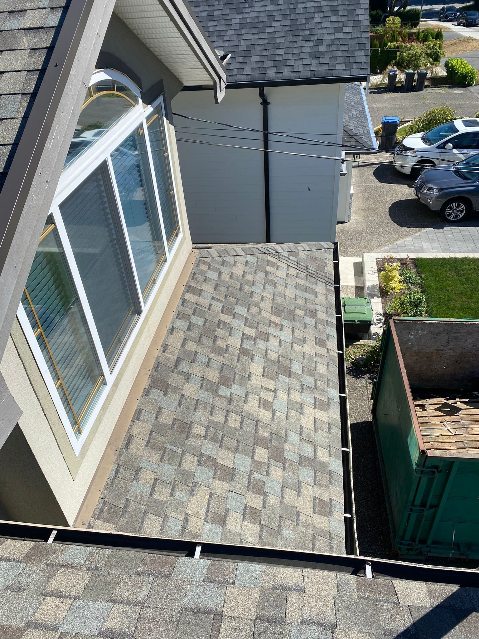 A high-angle view of a newly shingled residential roof, with a large green dumpster positioned in the driveway below.