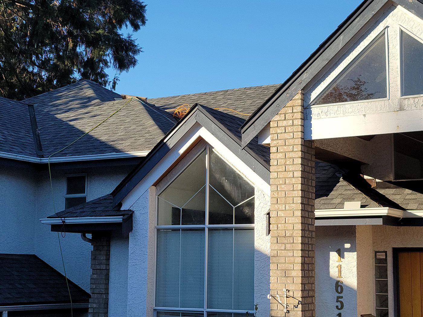 A house exterior featuring a brick chimney, a large window, and gray roof shingles under a clear blue sky.
