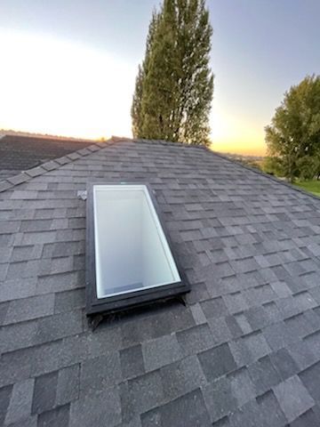 A rectangular skylight installed on a dark gray shingled roof under a clear sky at sunset.