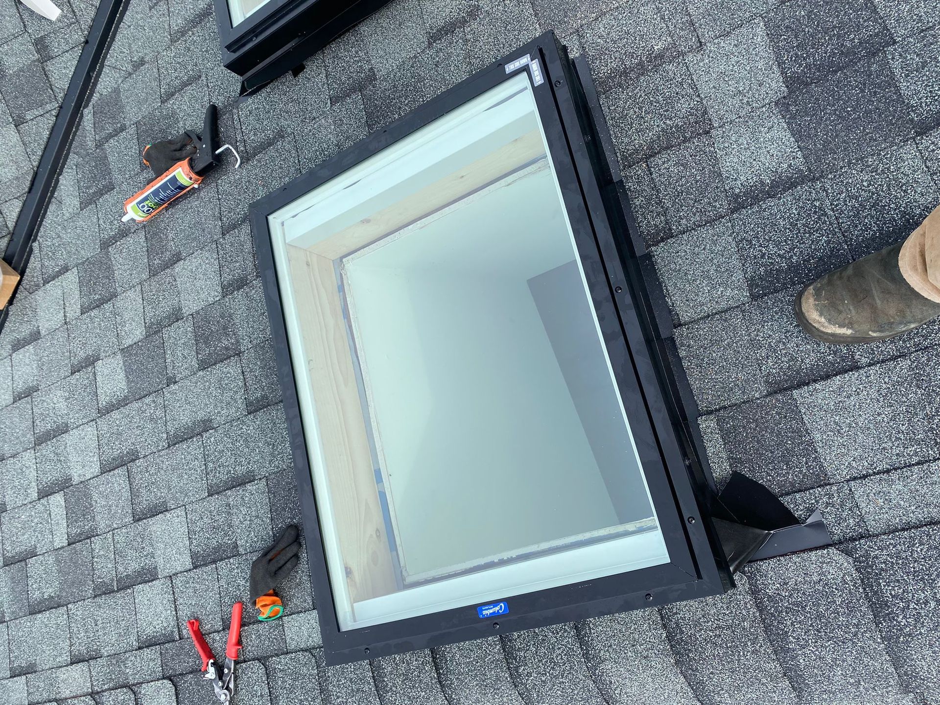 A black-framed skylight installed on a gray shingled roof, with construction tools visible nearby.