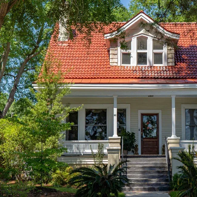 A two-story home with a reddish-orange tiled roof, white siding, and a front porch, nestled among green trees.