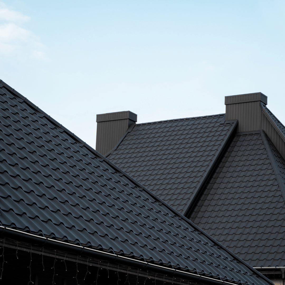 Close-up of a modern dark gray metal roof with two rectangular chimneys against a clear, pale blue sky.