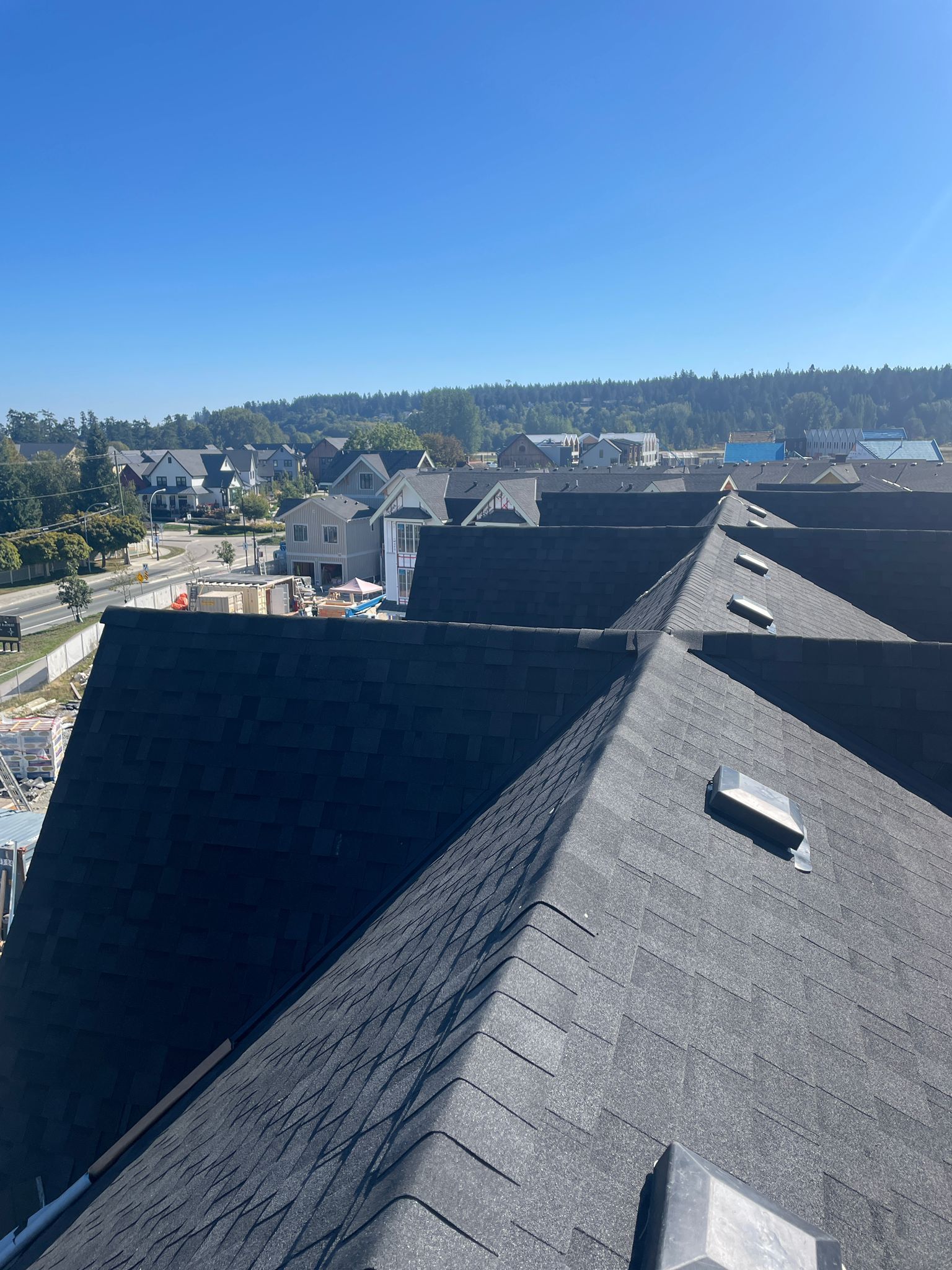 A view from a high vantage point overlooking dark shingled rooftops, with a neighborhood and forested hills in the distance.