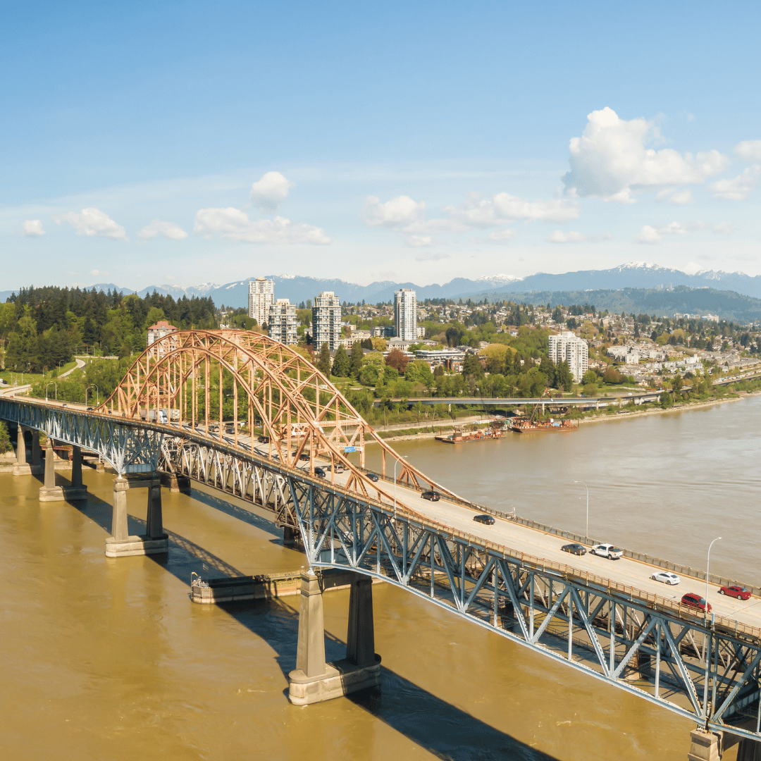 An aerial view of the Pattullo Bridge crossing a wide, muddy river with a cityscape and distant mountains in the background.