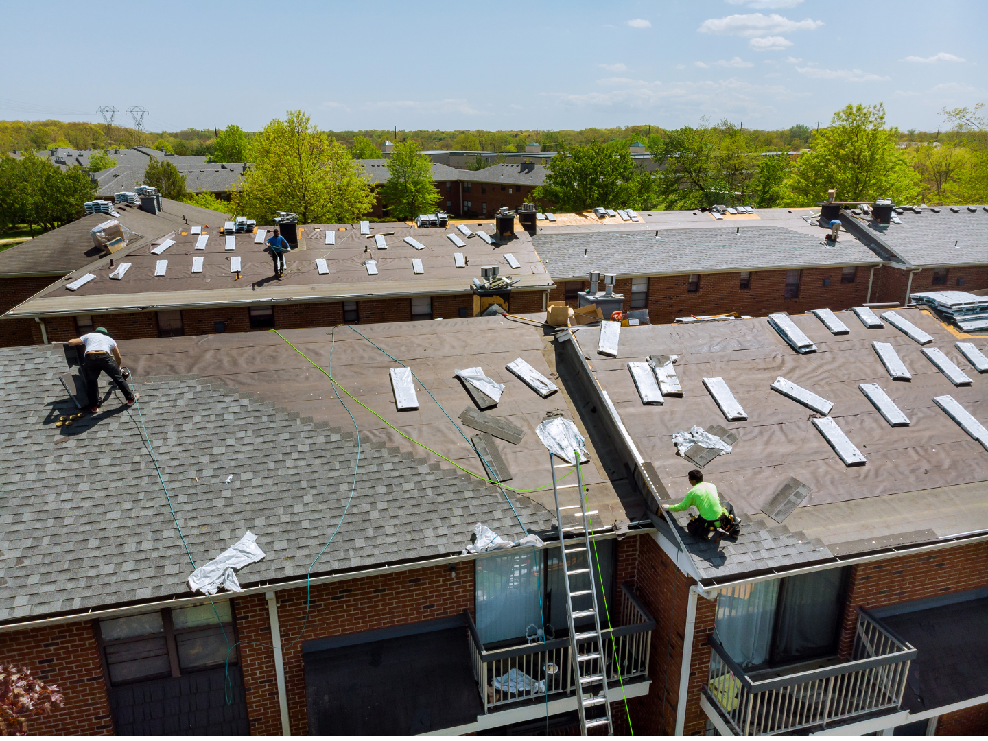 Workers in bright shirts are installing shingles on the flat roof of a multistory brick residential building.