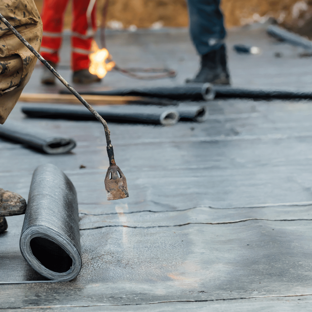 Workers use a propane torch to heat and install black bituminous roofing membrane on a flat surface.
