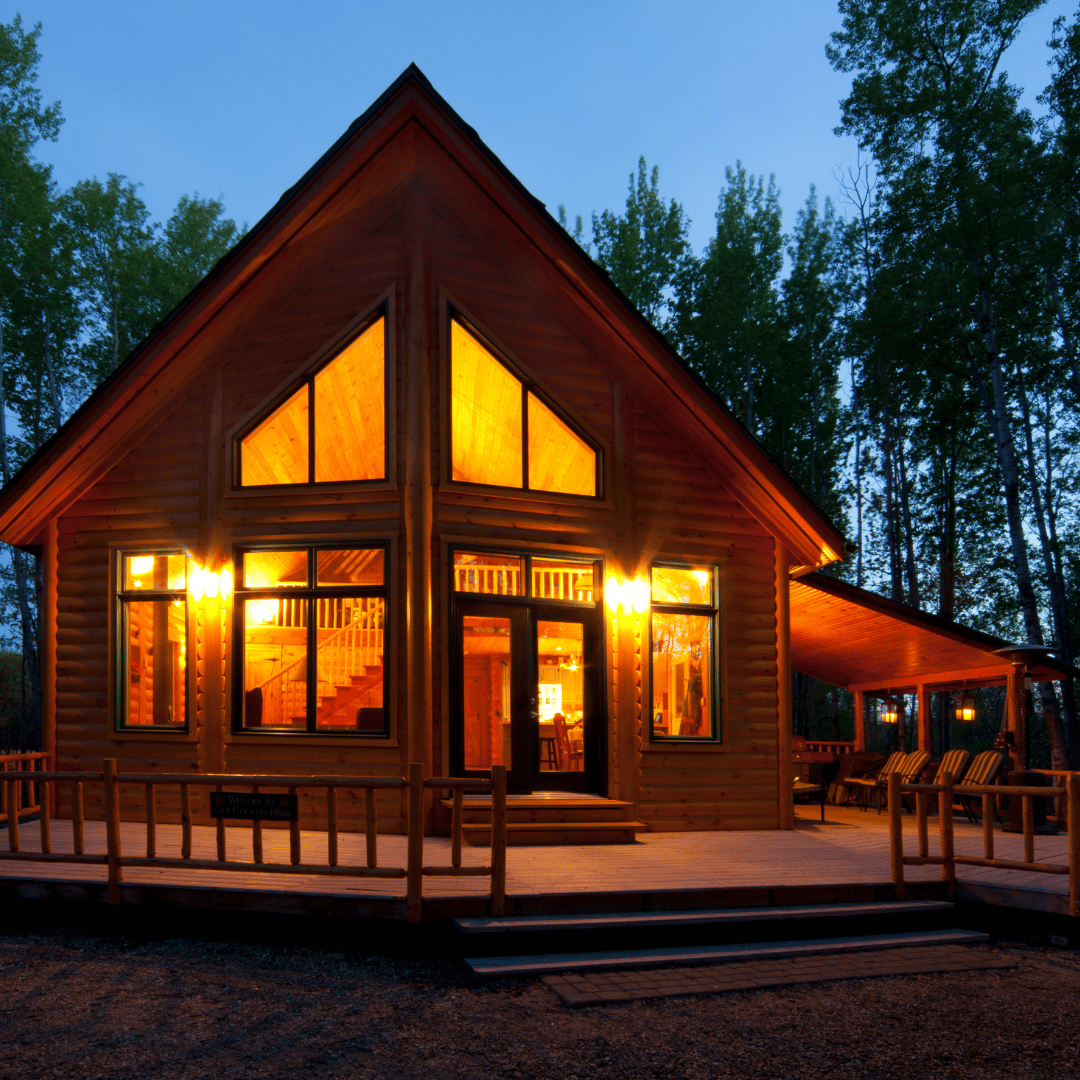 A warm, glowing wooden cabin at dusk, surrounded by trees with a porch and lit windows.