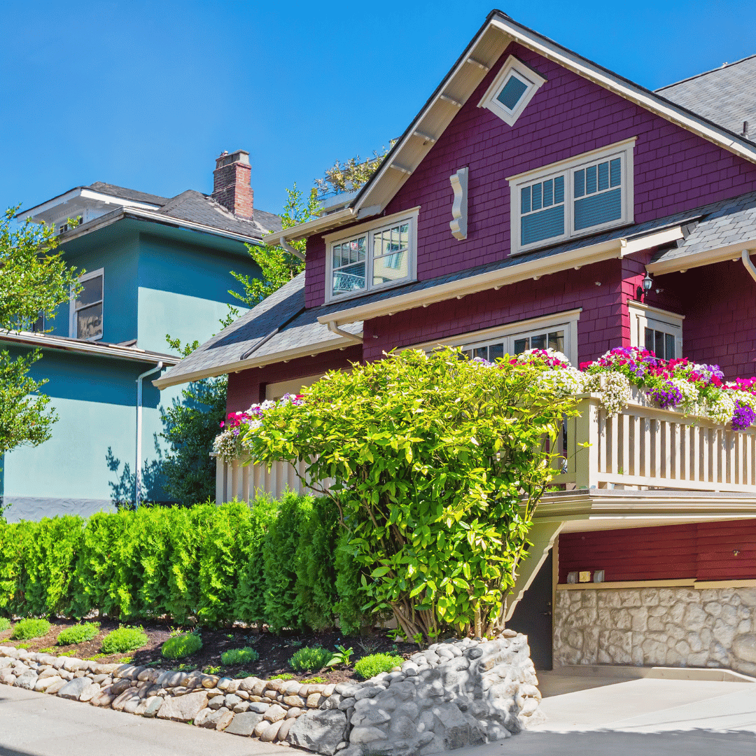 A bright purple house with a stone foundation, wooden balcony, and a green shrub garden next to a teal house.