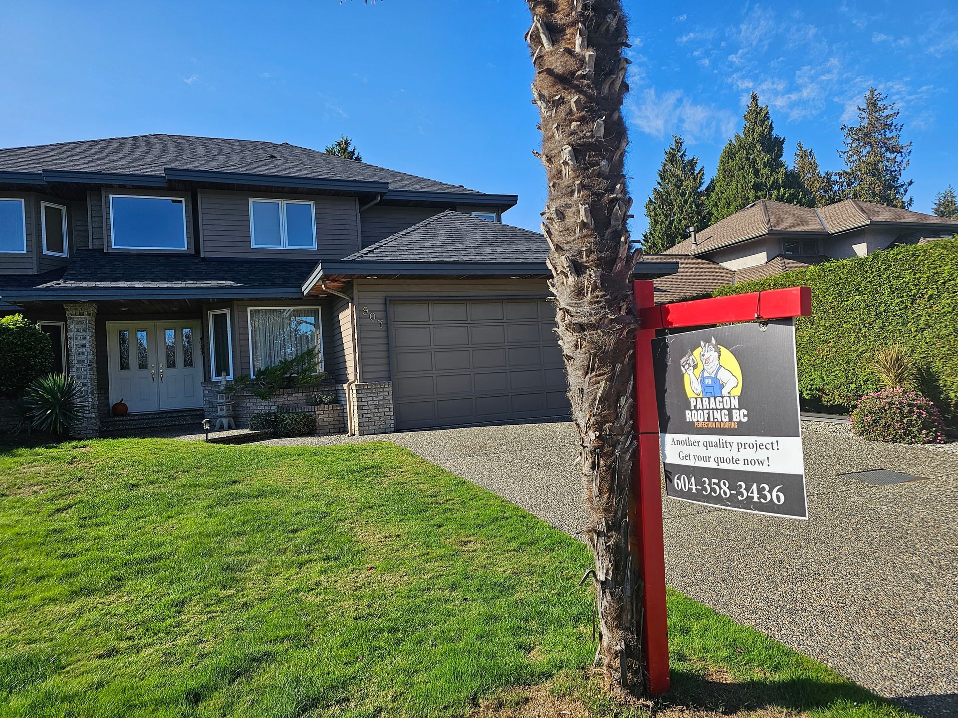 A brown two-story house with a green lawn and a real estate sign in the foreground under a clear blue sky.