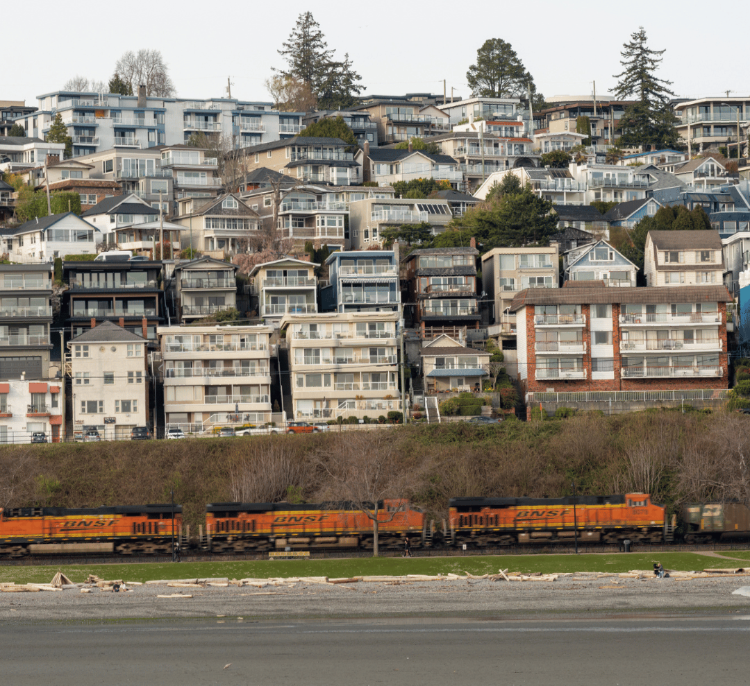 A freight train passes along a beach, with numerous houses densely clustered on the hillside above.