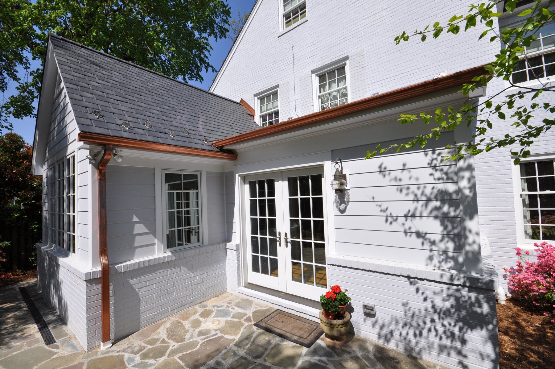 White house exterior with stone patio, French doors, and red flower pots in sunlight in Rockville