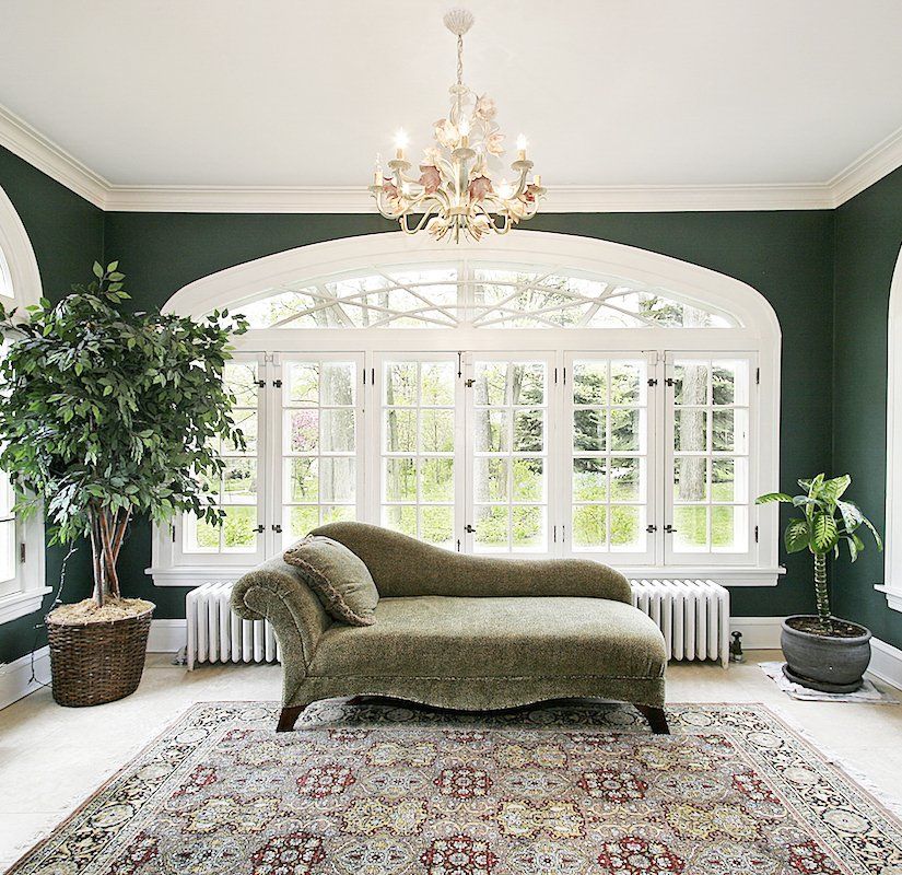 Elegant living room with green walls, arched windows, chaise lounge, chandelier, plants, and patterned rug in Montgomery Country MD