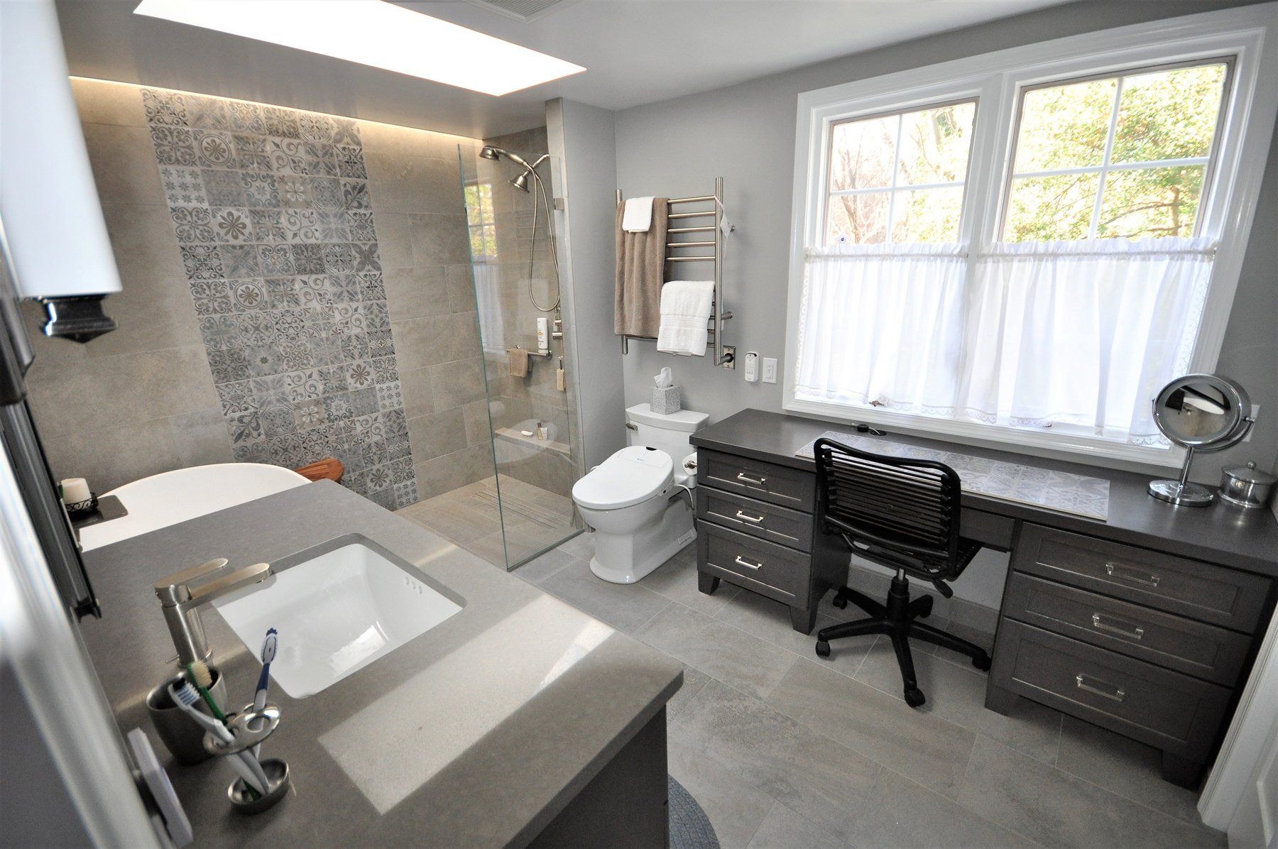 Modern gray bathroom with double vanity, toilet, tiled shower, and large window with desk chair underneath in Rockville