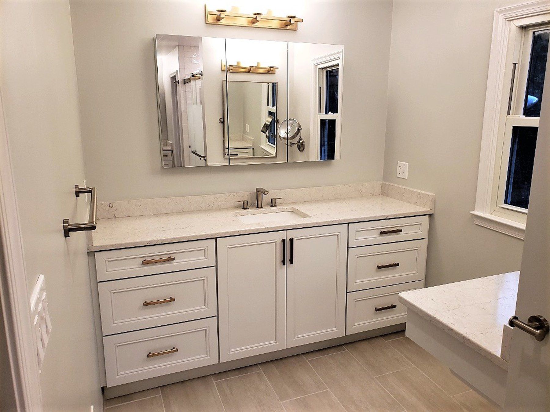 Bright bathroom with white double vanity, marble countertop, mirror, drawers, and bench by the window  in Rockville