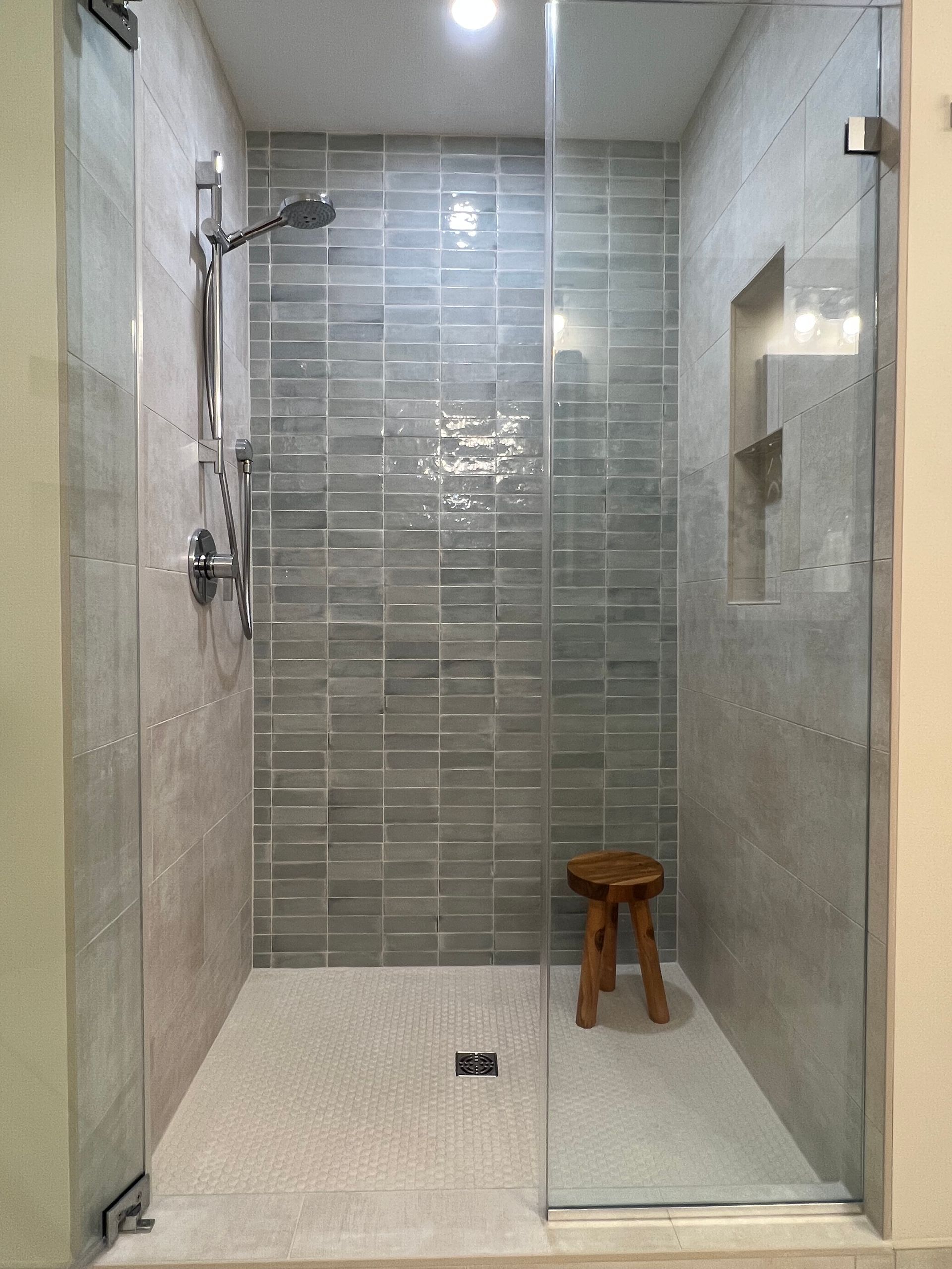 Modern glass shower stall with gray tile walls, white floor, and a small wooden stool  in Rockville