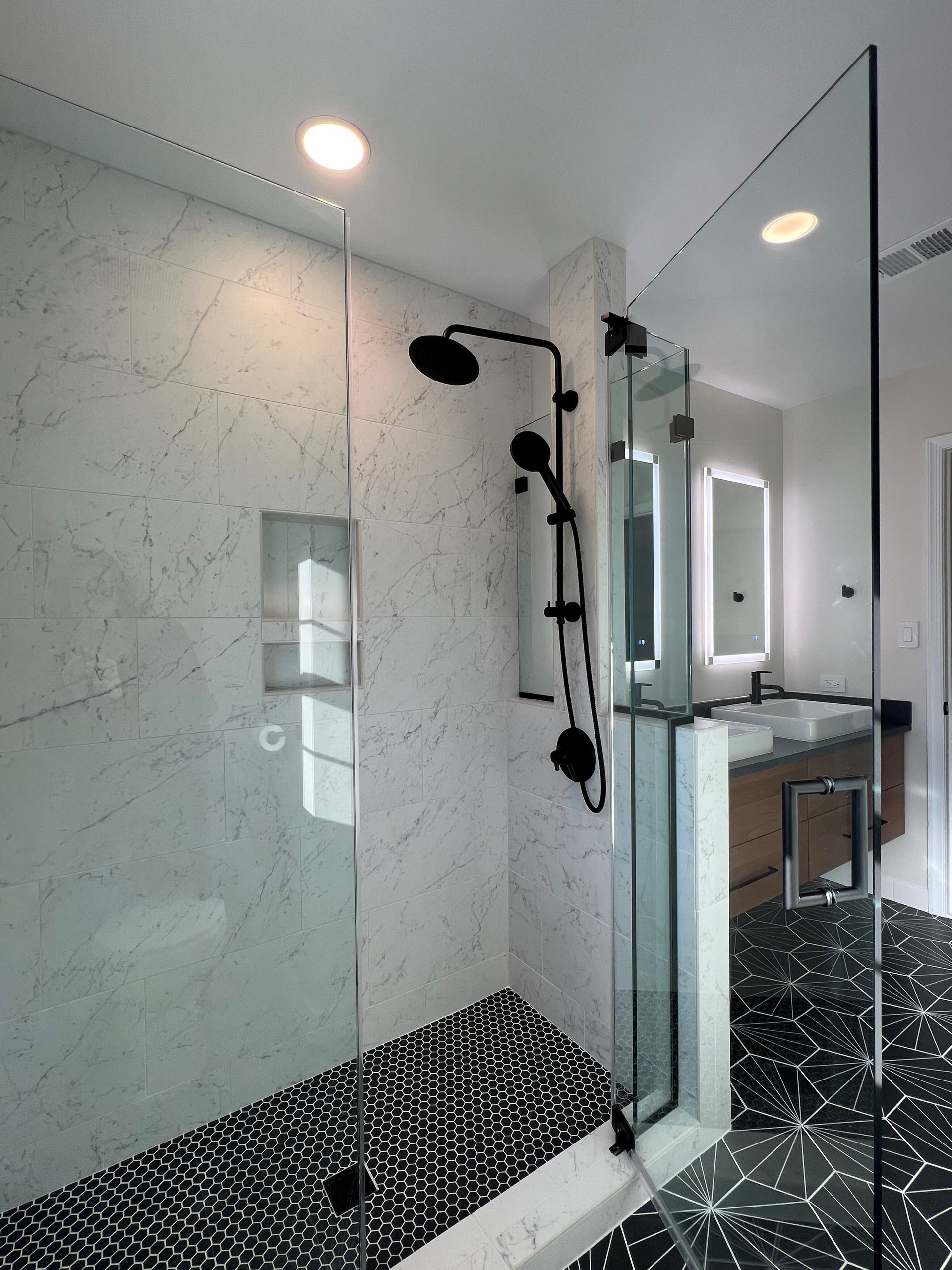 Modern shower with glass enclosure, black fixtures, and speckled black-and-white bench in a tiled bathroom  in Rockville