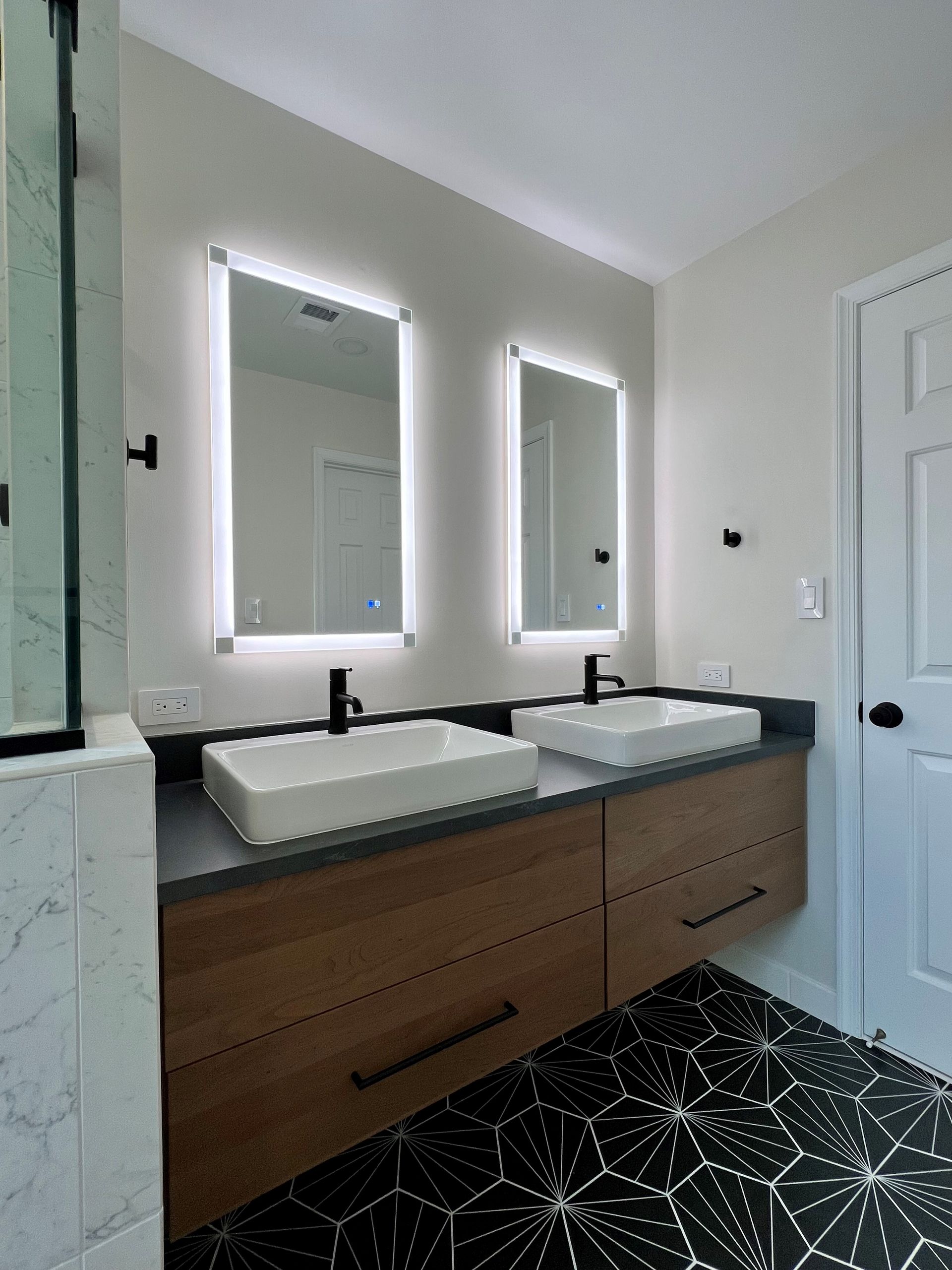 Modern bathroom vanity with dual sinks, lit mirrors, wood cabinets, and a black patterned rug  in Rockville