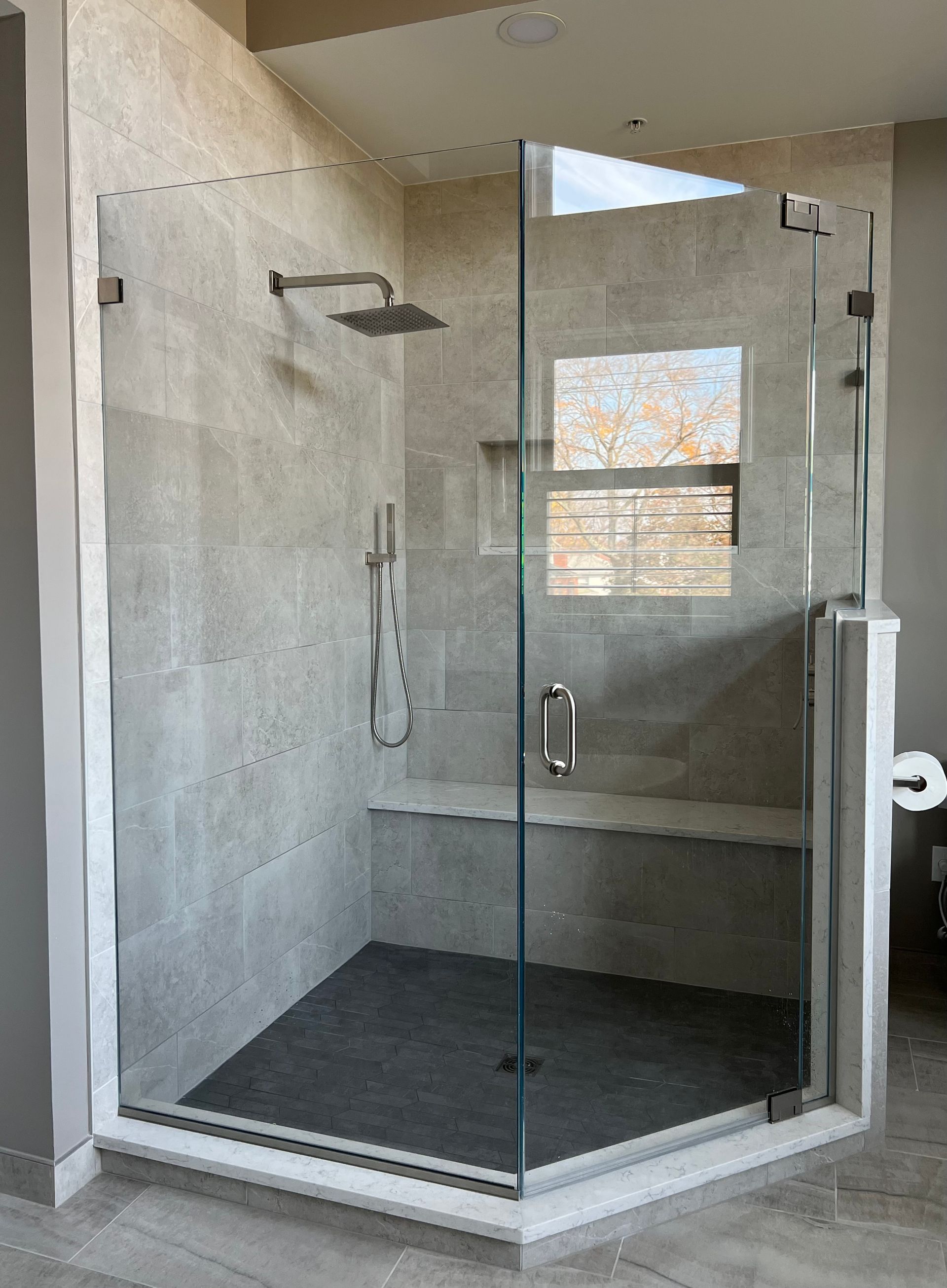 Glass walk-in shower with gray tile, black floor, and a built-in bench, viewed from the bathroom doorway  in Rockville