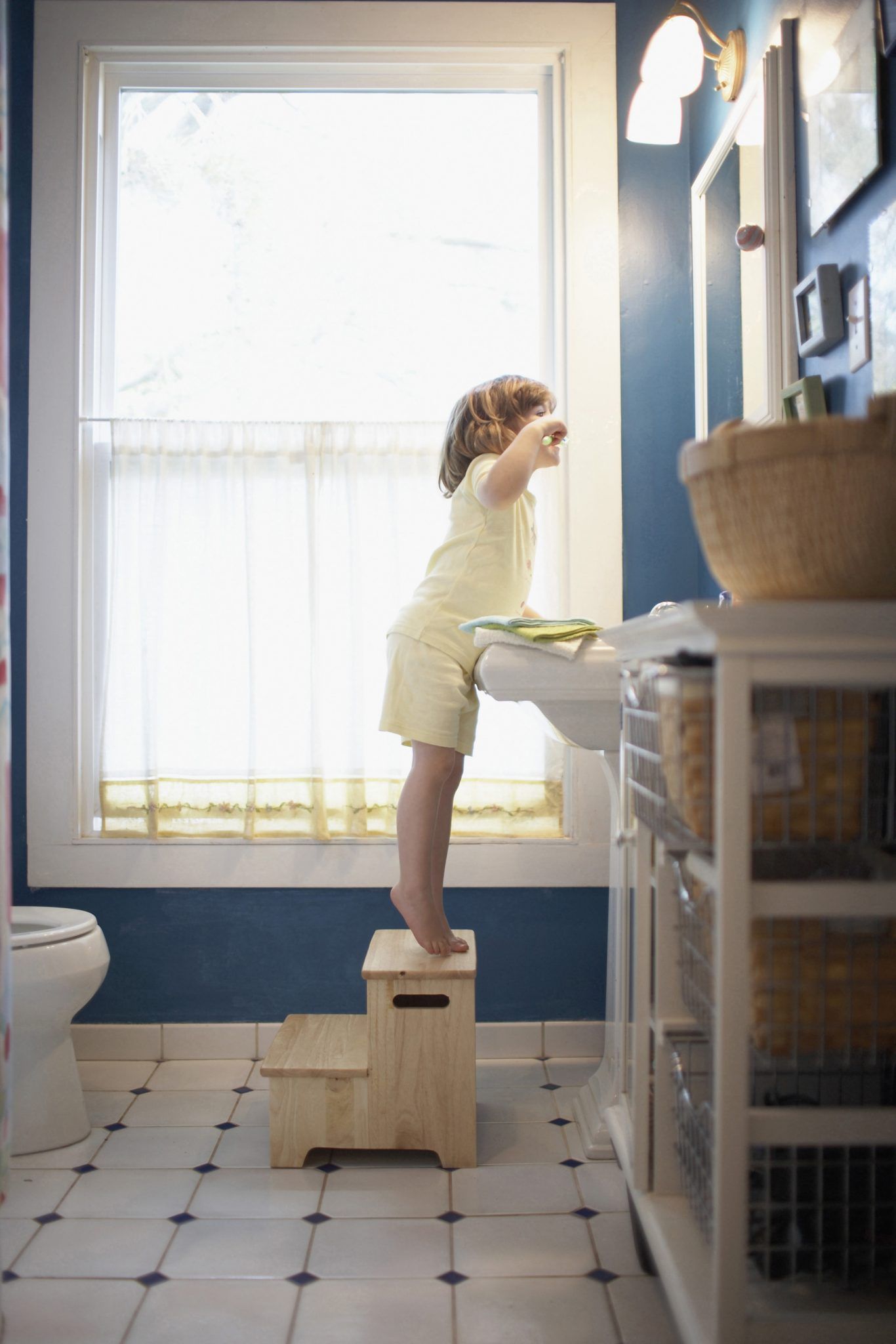 Child standing on a wooden step stool at a bathroom sink, reaching toward the mirror in Rockville