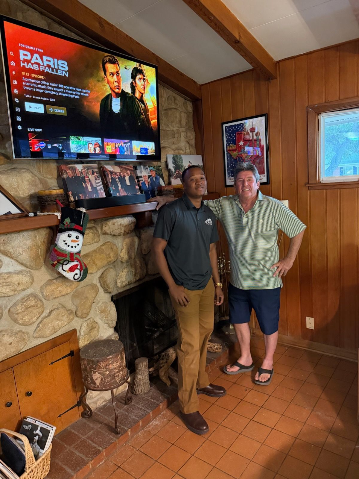 Two people standing by a fireplace in a wood-paneled living room under a wall-mounted TV.