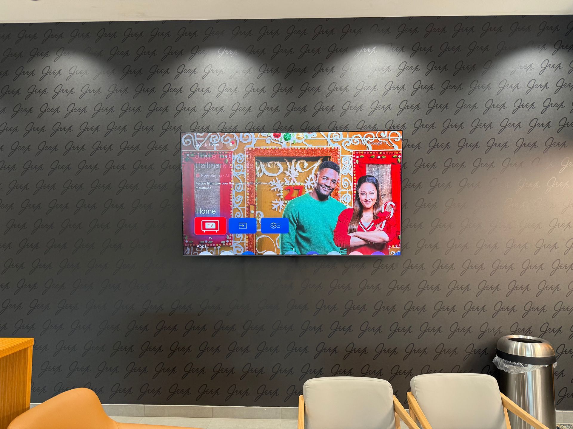 Wall-mounted TV on a dark wall in a lobby, showing two people with colorful graphics nearby.