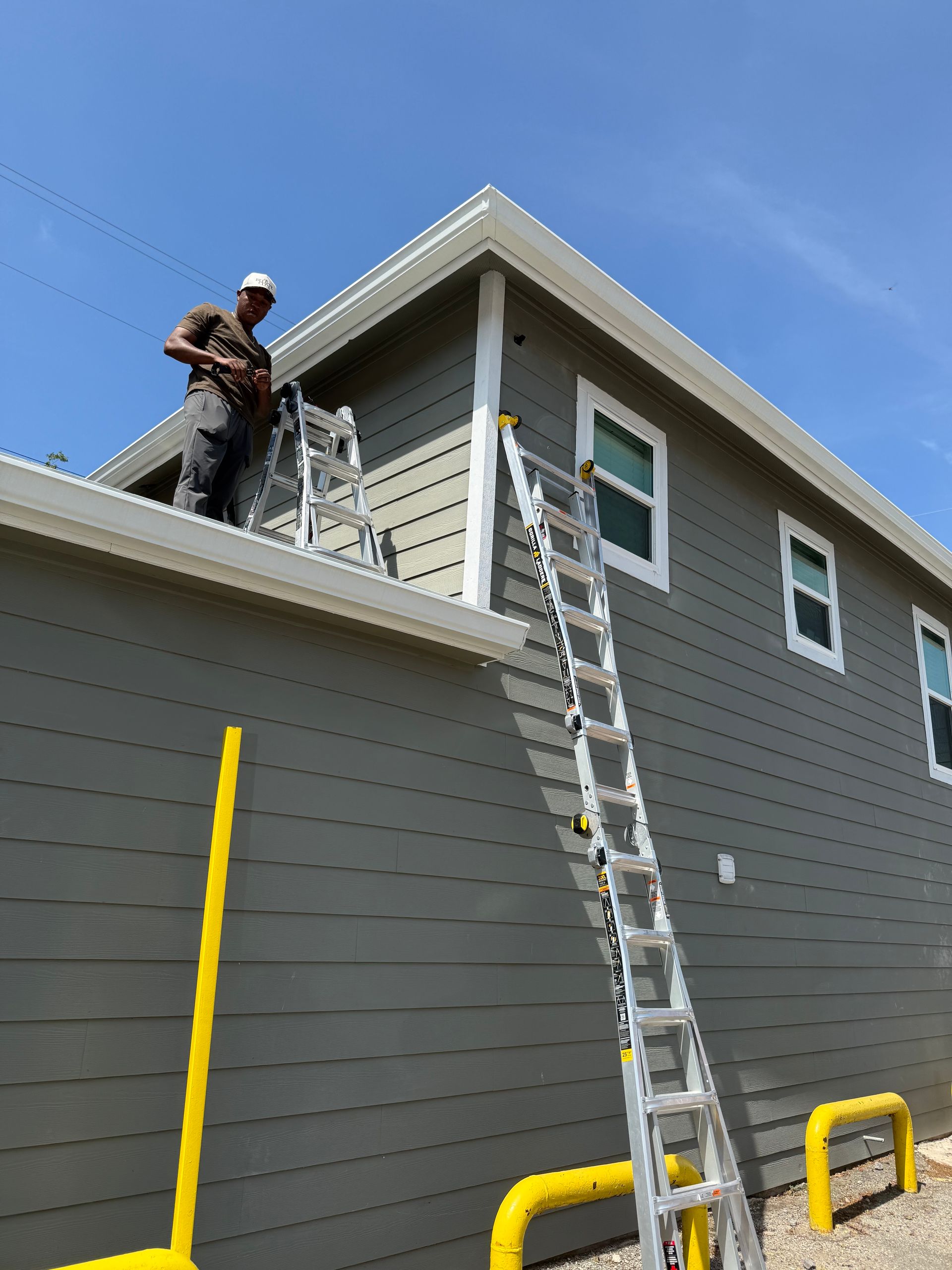 Workers on a roof beside a gray house, with ladders leaning against the eaves.