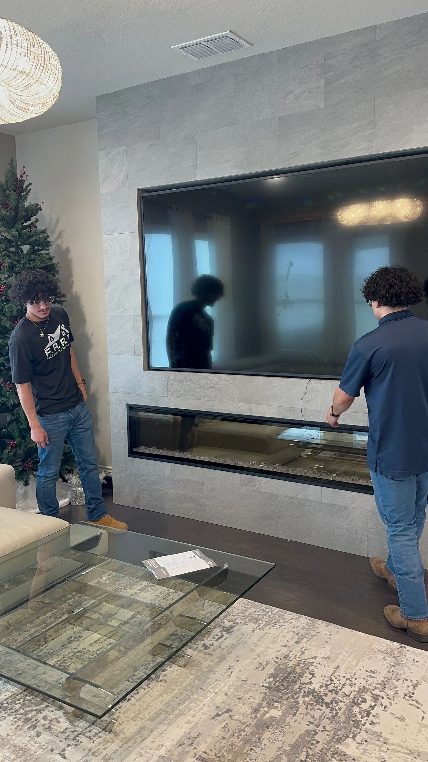 Two people standing beside a large wall-mounted TV in a modern living room, one adjusting the shelf below it