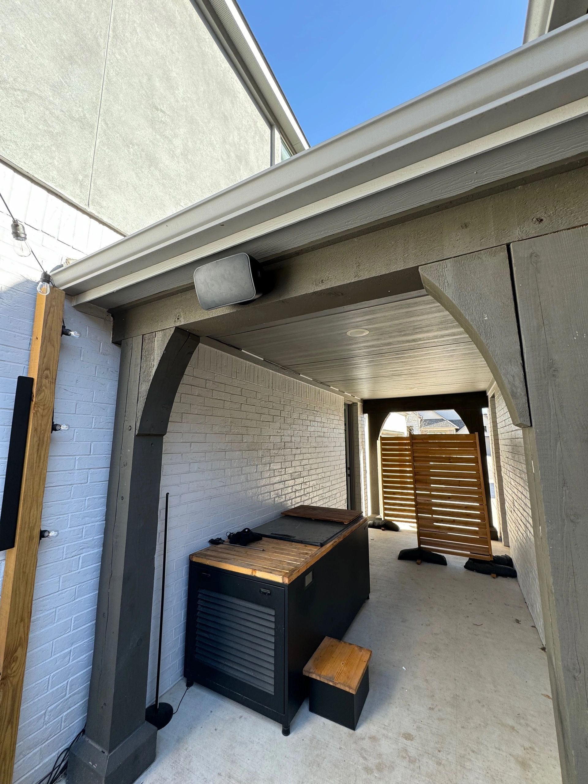 Covered outdoor patio with white walls, a black storage cabinet, and a wooden bench near a louvered door.