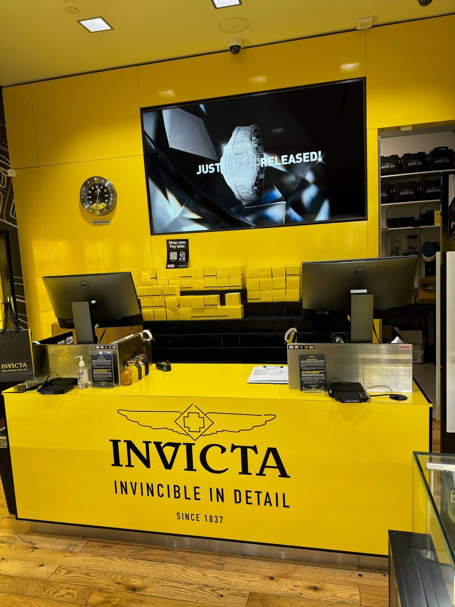 Yellow Invicta watch store counter with display screens and a branded desk in the foreground
