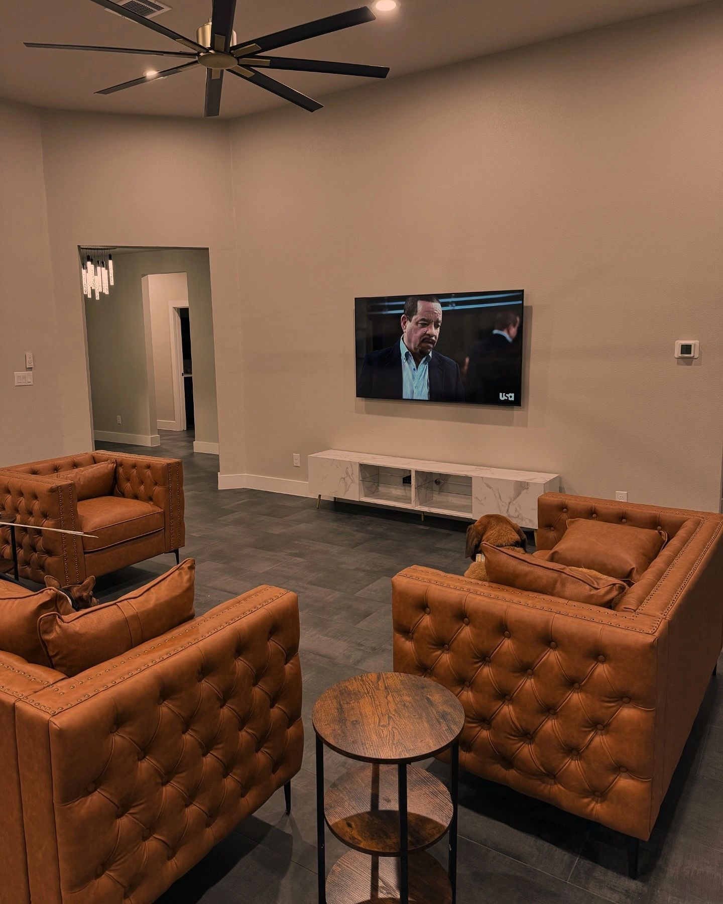 Modern lounge with tan tufted chairs, round table, wall TV, and gray carpet.