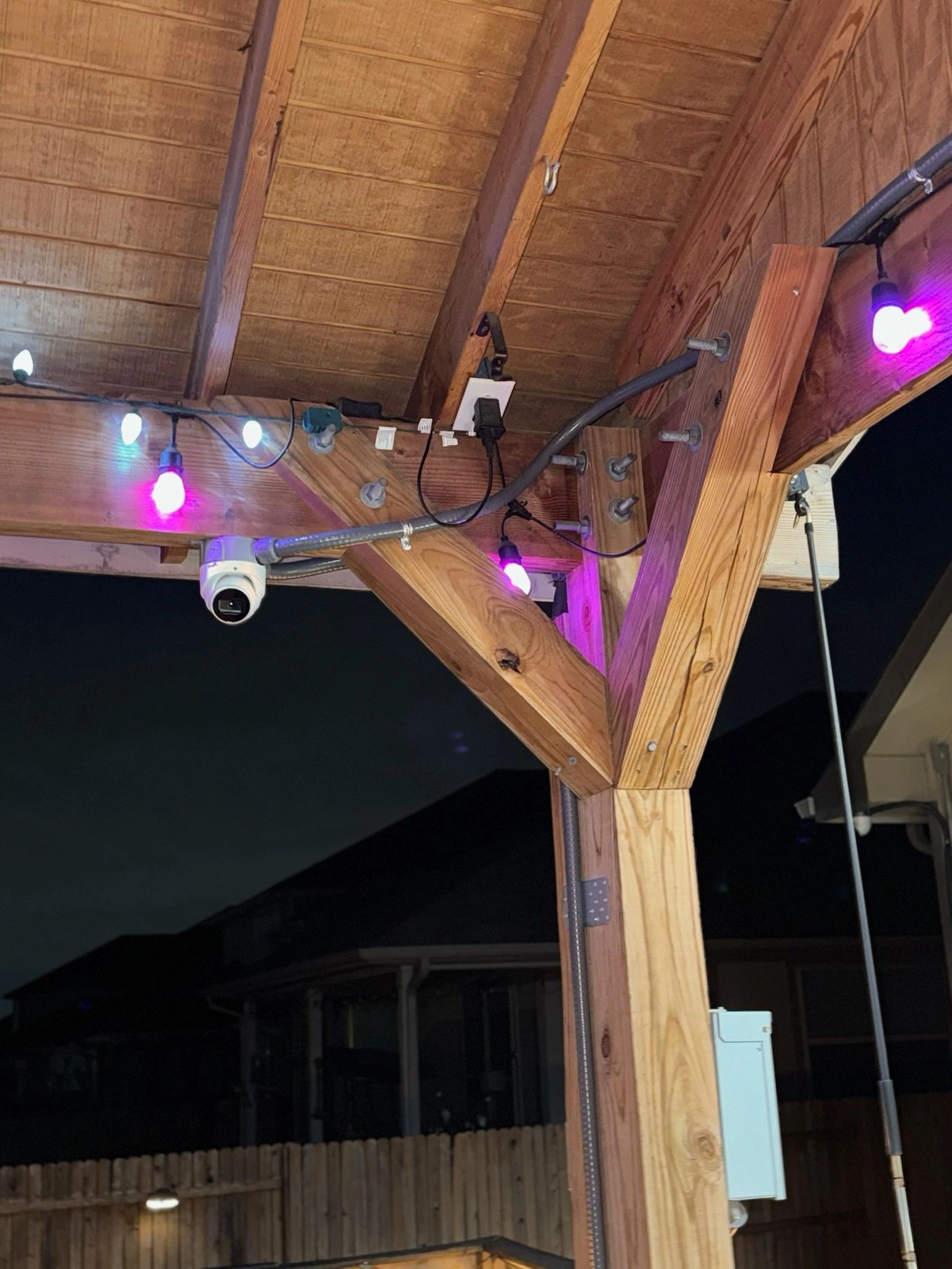 String lights and a security camera hang under a wooden patio roof at night.