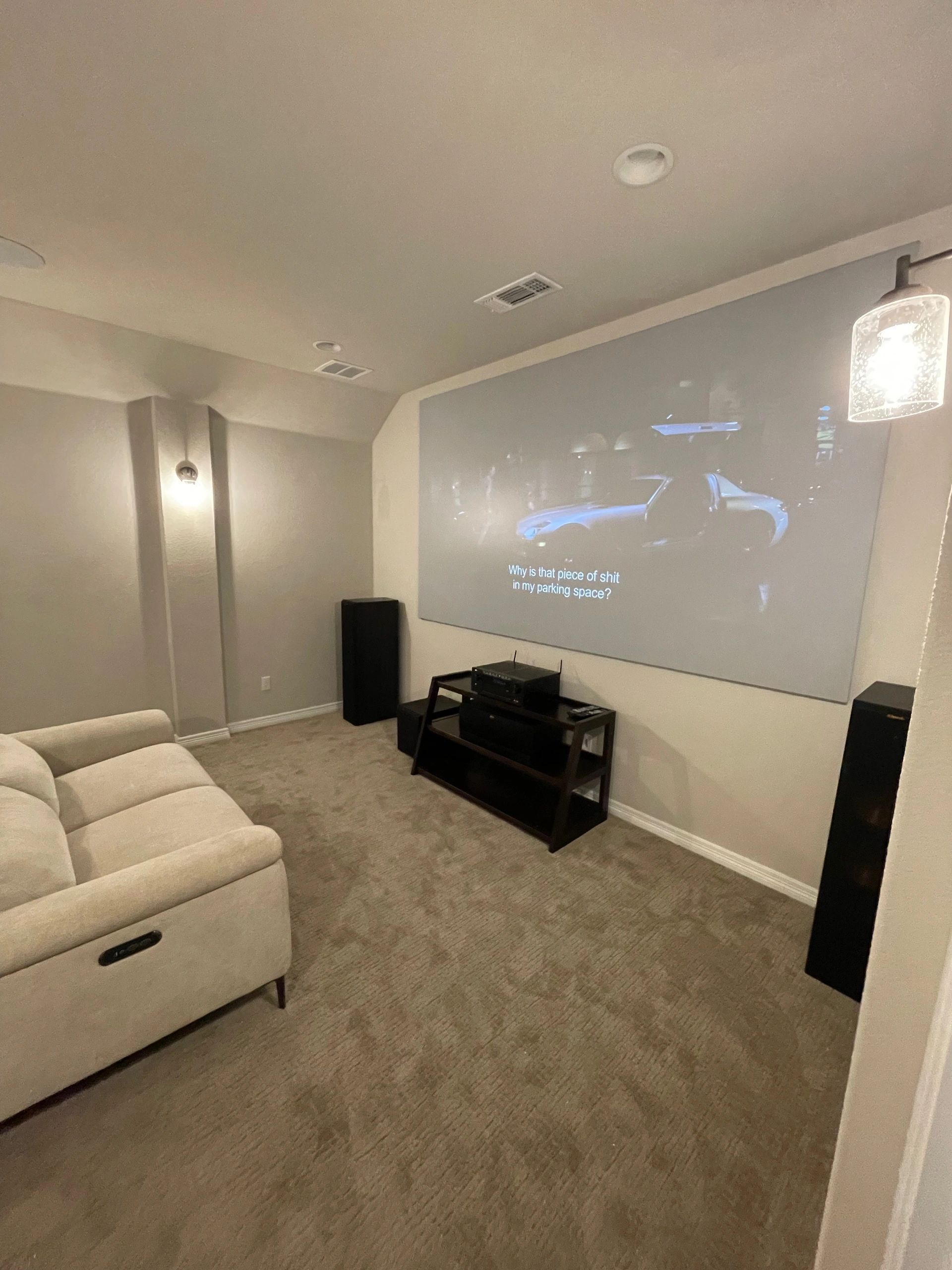 Cozy home theater with a beige sectional, projector screen, and black speakers in a carpeted room.