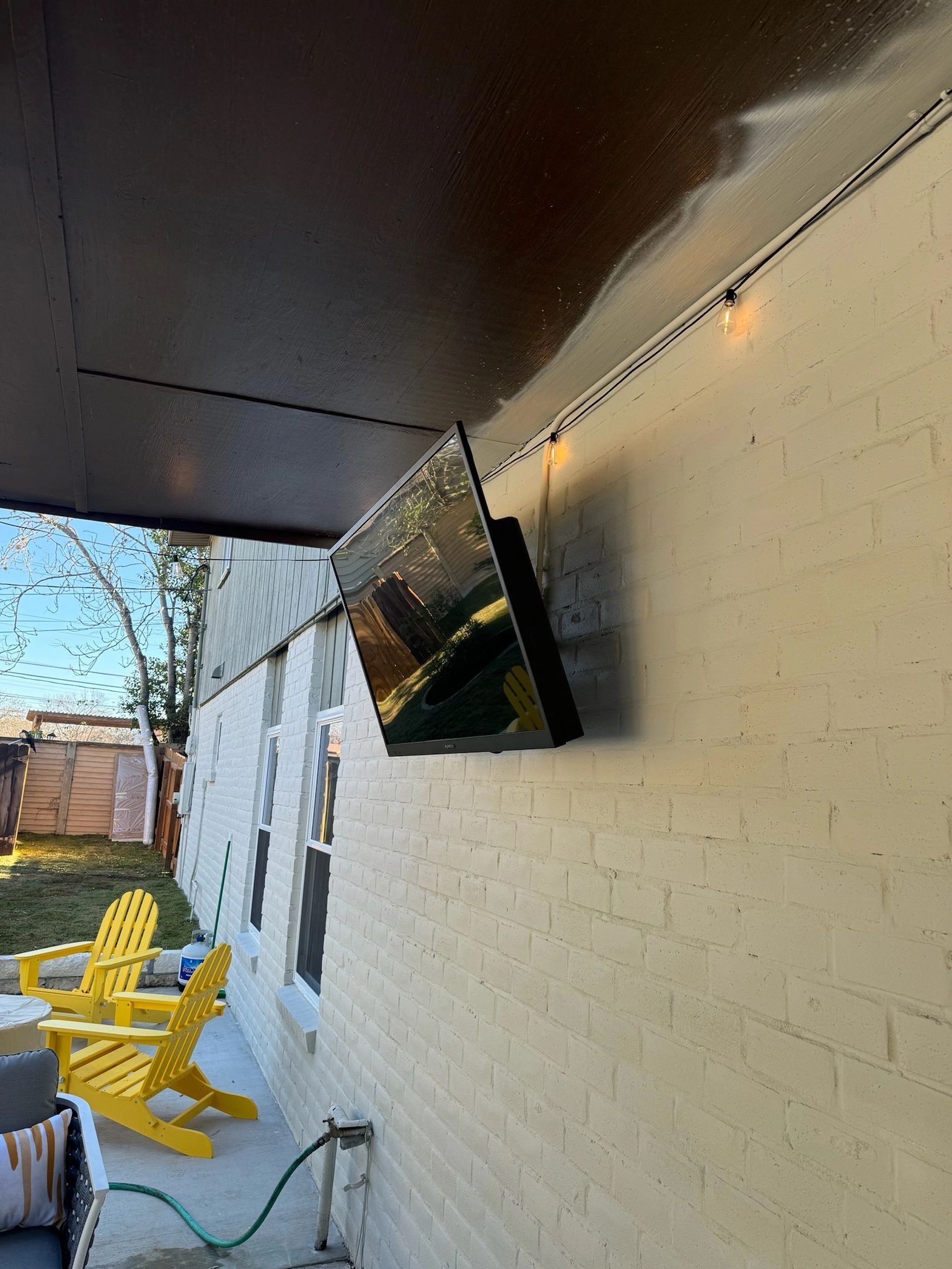 Outdoor patio with yellow chairs, white brick wall, and a wall-mounted TV under a dark overhang.