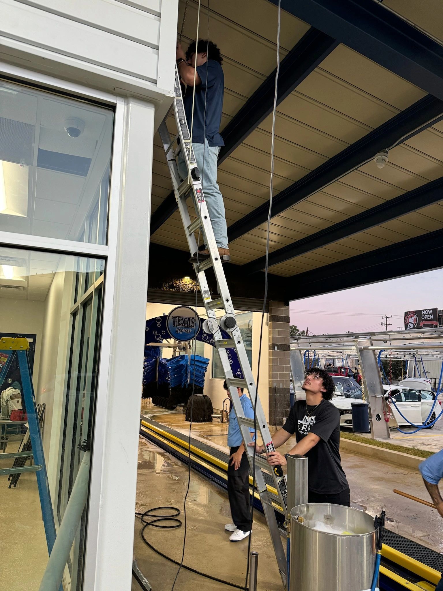 Person on a ladder working above a storefront while another steadies it at dusk under an awning