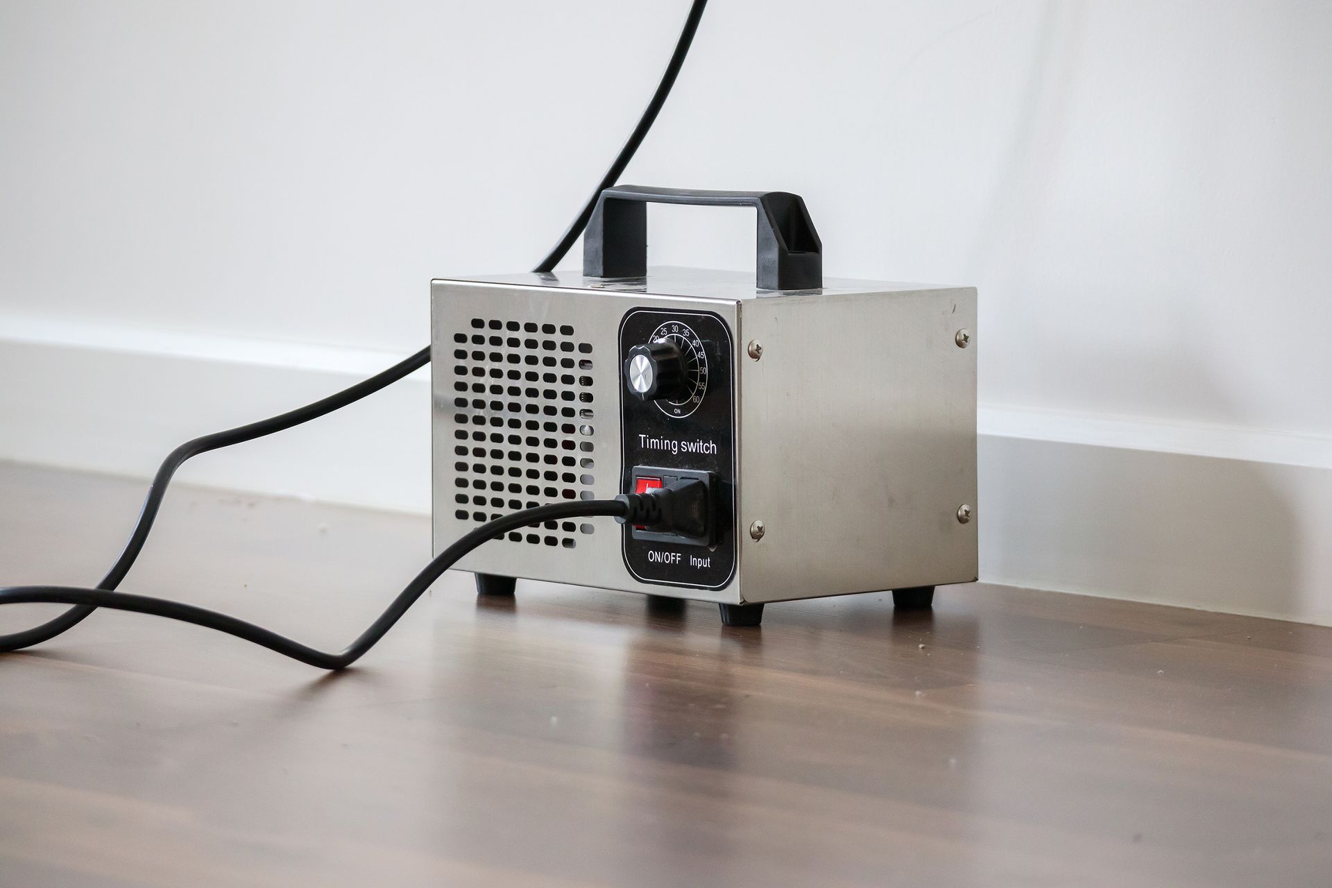 Small metal device with a handle and power cord on a wooden floor against a white wall.