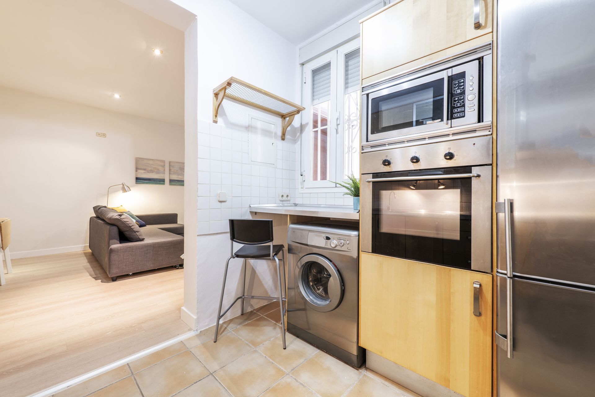 Compact kitchen with stainless steel appliances, washing machine, and stool opening into a living area.