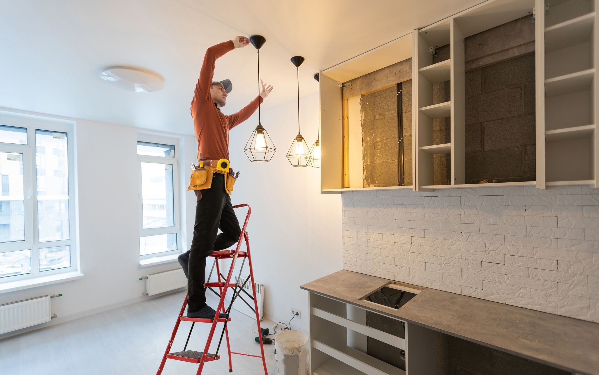 Person on a red ladder installing light fixtures in a bright, unfinished kitchen with cabinets open