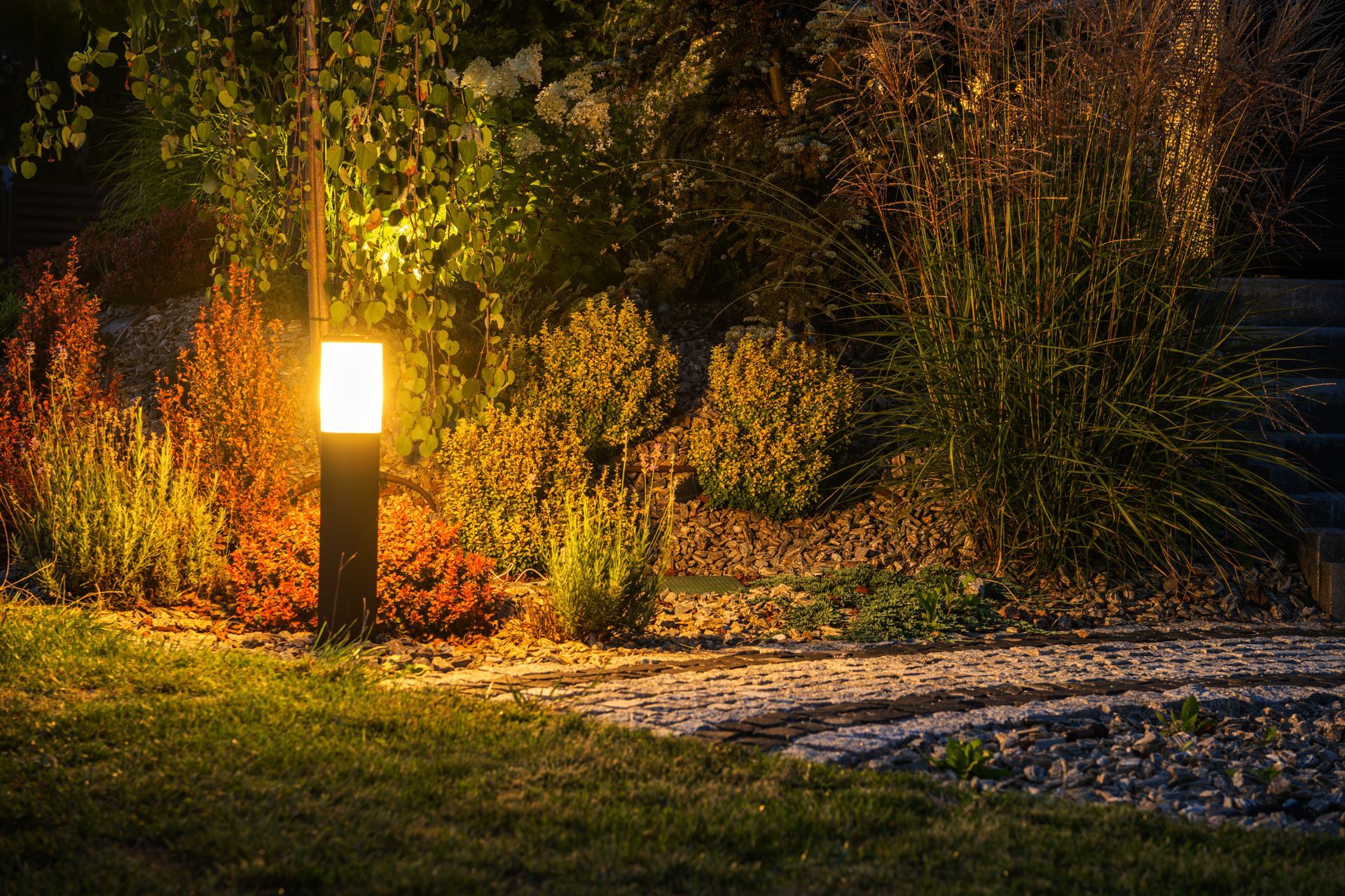 Warm-lit garden path with a glowing lantern beside shrubs and tall grasses at night