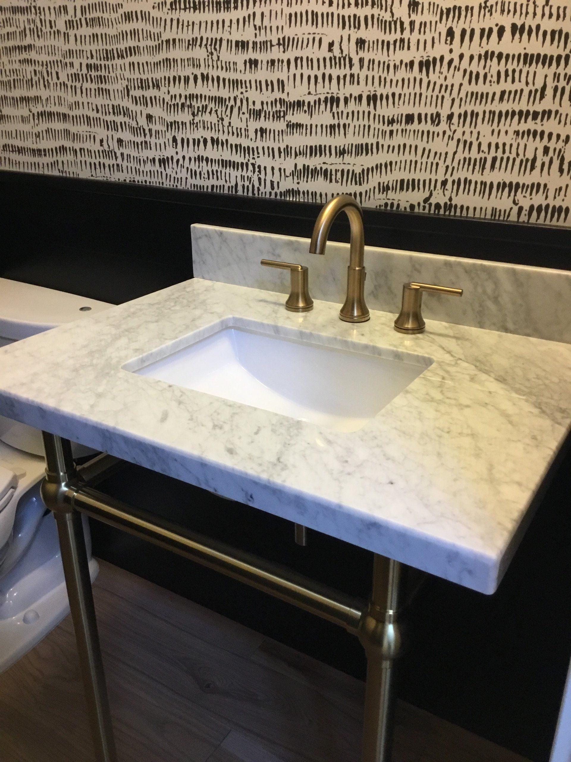Bathroom vanity with white countertop, under-mount sink, and brass faucet against patterned wall.