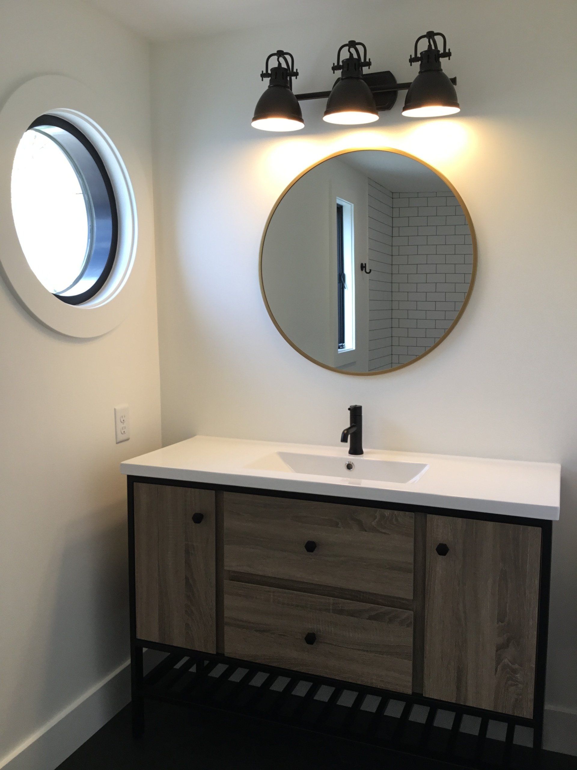Modern bathroom vanity with round mirror, black light fixture, and small round window.