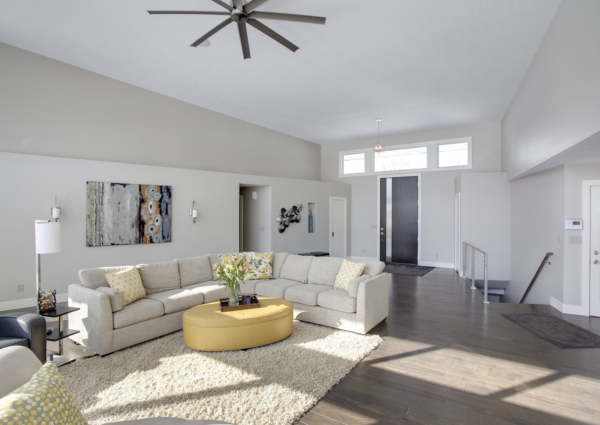 Bright modern living room with beige sectional sofa, round gold coffee table, rug, and vaulted ceiling with fan.